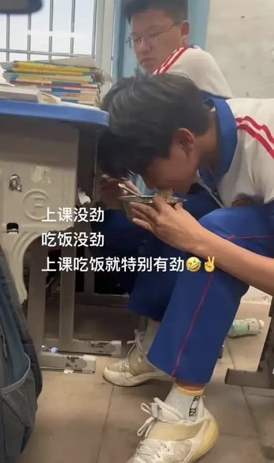 图片展示了两名穿着蓝白相间校服的学生在教室里的场景。一名学生正蹲在课桌下，低着头用勺子从碗里吃饭，姿态显得十分专注和投入。另一名学生则坐在座位上，平静地看着同伴的行为。课桌上堆放着一些书籍，教室环境看起来是典型的中学课堂。图片上的文字内容为："上课没劲 吃饭没劲 上课吃饭就特别有劲✌😋"，幽默地表达了学生在课堂上偷偷吃饭时反而精神十足的有趣现象，展现了校园生活中常见的调皮场景。