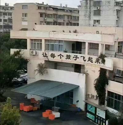 这是一张户外拍摄的现实场景照片，主体是一栋带有教育标语的建筑，建筑墙面上醒目地写着“让每个孩子飞起来”的字样。建筑外观略显老旧，墙面上生长着一些绿植，下方区域设有深色遮阳棚和橙色的休息座椅，背景是居民楼和茂密的绿植。这张图的趣味点在于标语的字面含义和建筑的位置结合，容易让人产生字面意义上的联想，形成搞笑的效果。