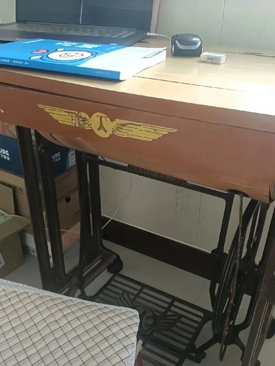 这张图片展示了一个将老式脚踩缝纫机改装为书桌的场景，上面放有课本、笔记本电脑和橡皮等学习用品。复古缝纫机和现代学习用品的组合产生强烈对比，带有一定的趣味和怀旧感。图中有明显的改造痕迹，暗示节俭利用资源，也可能带点自嘲或调侃的意味。没有明显双关、谐音梗或明显暗示成分。