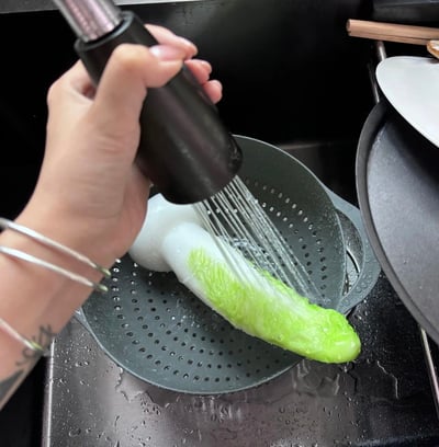 一只手握着黑色喷水器正在厨房水槽中冲洗绿色蔬菜（看起来像是生菜或卷心菜），蔬菜放在带有孔洞的滤水篮中，水流正从喷水器中喷出冲洗蔬菜表面，水槽周围可见一些厨具，整体场景展现了日常烹饪前的食材清洗过程。