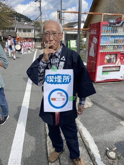 这是一张在日本街道拍摄的实拍趣味图，画面主体是一位戴眼镜的白发老年男性，他身穿日式传统外褂，内搭格纹衬衫，胸前用绿色胶带贴着一张写有日文“喫煙所”（即吸烟区）的告示，告示上还配有蓝色圆圈圈住的吸烟标识，老人正站在路边叼着香烟吸食，相当于把自己变成了移动吸烟区，创意十足充满幽默感。背景能看到路边的红色可口可乐自动贩卖机，远处的街道上还有穿着统一服饰、抬着神轿、敲着鼓的节庆游行队伍，周边是日式民居，看起来是当地举办民俗活动的场景。
