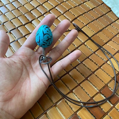 一只手握着一个蓝色蛋形吊坠，吊坠上有黑色图案。吊坠通过金属环连接到棕色绳子上，背景是木质网格状表面。