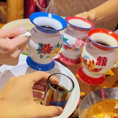 这是在热闹的聚餐饭桌上，几只被设计成老式搪瓷痰盂样式的杯子被用来盛装深色饮品，还有一只普通小玻璃杯也盛着同款饮品，几只手分别拿着杯子，似乎正在碰杯。杯子上绘制着传统的牡丹、鸳鸯纹样，其中两个杯子印有红色的“囍”字，将日常的餐具做成这种复古又搞怪的造型，和聚餐喝酒的场景形成强烈反差，为聚会增添了满满的欢乐趣味，没有双关、谐音梗或暗示内容。