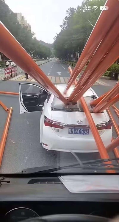 图片显示一辆白色丰田轿车（车牌号：粤M·6D533）在城市道路上发生意外，多根橙色管道从车辆前方贯穿车顶，管道末端向四周散开。车辆右侧 rear车门呈打开状态，周围可见道路施工隔离栏和行人和车辆，背景为绿树和建筑物。场景疑似因运输或施工中的管道掉落导致的交通事故，画面具有较强的视觉冲击力。