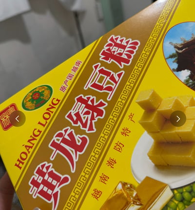 图片展示了一盒越南产的黄龙绿豆糕包装，主体为黄色和金色配色，正面印有红色大字“黄龙绿豆糕”，左上角标注“原产国：越南”及品牌名“HOANG LONG”，右侧有绿豆糕切块堆叠的实物图和古建筑图案。结合标题“三年前吃了一块, 噎到现在还没吞下去”，通过夸张手法幽默吐槽该绿豆糕口感干燥，容易噎人，形成搞笑效果。