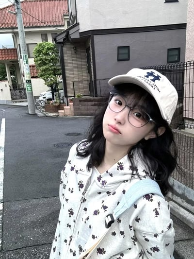 一位年轻女性站在日本的街道上，背景是典型的日式住宅建筑。她戴着一副透明框架眼镜，一顶印有蓝色字母'H'和文字的白色帽子，以及一件带有紫色小花图案的白色连帽衫。她的头发是黑色的，长度及肩，微微卷曲。女孩微微歪着头，表情平静地看向镜头，背景中可以看到铺着红色瓦片的屋顶、电线杆、自行车和一些绿植，营造出一种宁静的日系街道氛围。