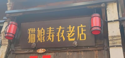 这是一张实拍的老街店铺招牌照片，深棕色的木质招牌上，用醒目的立体黄色字体写着「猫娘寿衣老店」，招牌左右两侧悬挂着带有中式纹样的复古红色灯笼，招牌下方的门梁处还有「老街25号」的棕色门牌号，整体呈现出古旧的传统店铺门面风格。店名中「猫娘」这个偏软萌的称呼和「寿衣老店」的严肃丧葬属性形成强烈反差，容易让人产生新奇感。
