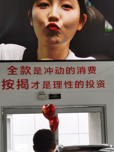 这是一张汽车4S店内的实拍图：上方屏幕播放着嘟嘴的女性画面，下方白墙上贴着醒目的红色营销标语「全款是冲动的消费，按揭才是理性的投资」，画面下方可见一个小孩的背影和印有五星红旗图案的爱心气球。这是商家为引导顾客选择贷款购车而设置的宣传内容，没有双关、谐音梗或暗示内容。