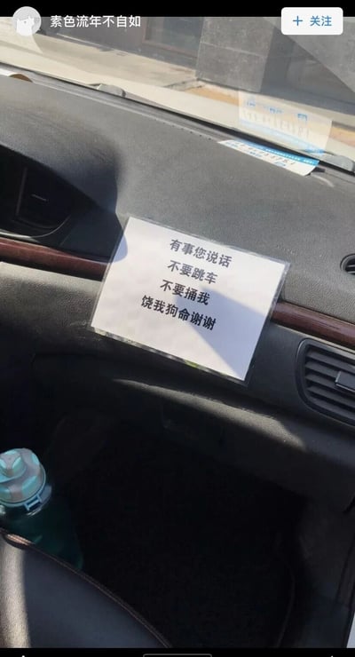 这是一张社交平台的截图，画面主体是汽车副驾驶前方的中控区域，这里贴着一张过塑的白色提示纸条，纸条上用黑色字体写着“有事您说话 不要跳车 不要捅我 饶我狗命谢谢”，以夸张、自嘲的语气，表达了网约车司机对遇到极端乘客的担忧，用搞笑的方式传递出自身的诉求。画面左下角能看到一个带图案的水杯，图片顶部显示发布者的用户名“素色流年不自如”以及“+关注”按钮，整体风格诙谐幽默，是对行业内极端事件的调侃。