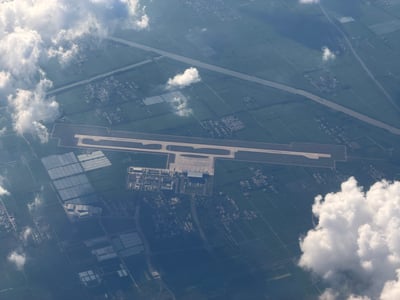 这是一张从巡航阶段民航客机舷窗向下拍摄的日间高空航拍图，当天天空分布有零散积云。画面核心是一座建在开阔平原上的小型民用机场：机场设一条近东西走向的长跑道，跑道南侧配套平行滑行道与联络道，停机坪、长条形现代航站楼及后勤工作区全部布置在跑道南侧，整座机场由围界圈合，轮廓清晰规整。机场四周是被田埂、道路分割成规则方块的绿色农田，田间散落多处小型村落聚居点；机场北侧约3-4公里处有一条水面宽阔、河道笔直、东西向横贯整个视野的河流；机场西南侧分布着数片矩阵排列、表面反光的连片设施，形态符合农业温室大棚或光伏发电站特征。画面左上角和右下角有大团蓬松的白色积云，部分地面被薄云阴影覆盖，视野开阔通透。结合拍摄者提供的航班信息（广州白云经停武夷山飞往青岛胶东的SC4678航班，拍摄时间为8月25日16:39），对照地理信息可判断该机场为原济宁曲阜机场（嘉祥场址）：其单条东西向跑道、北侧东西走向的洙赵新河、周边鲁西平原农田地貌、西南侧连片温室的特征均与画面完全吻合，且该场址正处于武夷山飞往青岛的航线途经范围内。