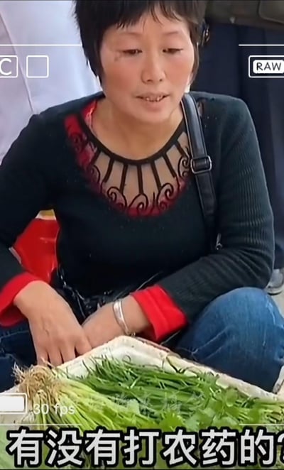 图片中是一位坐在地上卖菜的中年阿姨，她穿着黑色带红色装饰的毛衣和蓝色牛仔裤，面前摆放着新鲜的绿色蔬菜。画面带有类似相机拍摄的界面元素，底部有字幕"有没有打农药的?"，看起来像是一段日常对话的视频截图。