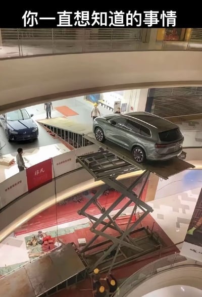这是一张从商场高层向下拍摄的实拍图，顶部配有文字“你一直想知道的事情”，画面呈现了商场展车的运输过程：工作人员操作剪式升降台将一台浅绿色SUV抬升到与一层展区坡道齐平的高度，准备将车辆驶到一层展区，坡道旁还停放着另一台蓝色展车，有数名工作人员在现场配合调度作业，解答了大众好奇的“商场里的展车是如何进入展位”的疑问。
