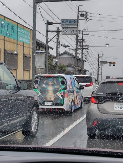 雨天的日本街道上，一辆汽车的后部被完整的动漫角色贴纸覆盖，角色为蓝发双马尾少女，穿着比基尼，双手举起。周围有其他车辆行驶，背景可见挂着日文招牌的建筑物和电线杆，包括"法務局"（法务局）和"メター21"等指示牌。车窗上的雨滴清晰可见，营造出真实的雨天氛围。
