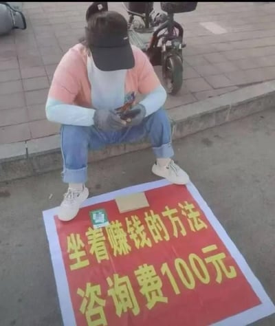 图片中一名戴着黑色帽子和口罩的人蹲坐在路边，身穿粉色开衫和蓝色牛仔裤，脚踩白色运动鞋，正低头看着手机。其面前铺着一块红色广告牌，上面用黄色大字写着“坐着赚钱的方法 咨询费100元”。背景可见砖铺人行道和一辆停放的电动 scooter，整体场景呈现出一种街头营销的幽默画面。该场景通过“坐着赚钱”的方法本身就是坐着收费咨询，形成一种循环讽刺的效果，暗指所谓的“赚钱方法”可能并无实际内容，仅为噱头。