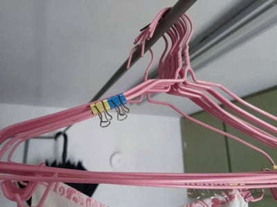 这是一张向上仰拍的居家场景实拍图，深灰色晾衣杆上挂着十余只统一的粉色浸塑衣架，其中部分相邻的衣架杆身被黄色、蓝色的长尾夹夹住固定，金属夹口朝下悬挂，衣架下方还能看到部分被悬挂的带有粉色印花的织物，背景可见白色墙面与浅色柜体，整体是十分生活化的衣物收纳晾晒场景。