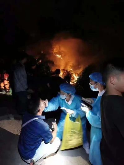夜晚的户外场景中，背景是熊熊燃烧的山火，火光将夜空映得通红，火势看起来较为凶猛。前景处，两位身着蓝色防护服、佩戴蓝色防护帽与口罩的医护人员，正专注地为一名蹲着的男子进行核酸采样，男子手持手机似乎在辅助照明。周围还有其他围观或待命的人员，一边是紧急的山火情势，一边是按流程推进的防疫采样工作，两种截然不同的紧急场景交织，形成了极具冲击力的画面，图中无任何双关、谐音梗或暗示类内容。