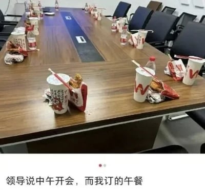 该图为图文混排的实拍梗图，上半部分展示了公司的会议长桌，每一个参会座位前都摆放了一份华莱士快餐套餐与瓶装矿泉水，还有拆开的食品包装散落在桌面上；下半部分配文为“领导说中午开会，而我订的午餐”，营造出本应严肃的会议场景被全员带午餐参会的行为消解的反差感，调侃了公司占用员工午休午餐时间开会的不合理职场现象。