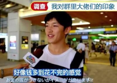 图片为一段街头采访的视频截图，场景可能在商场或车站等公共场所，背景有模糊的行人和店铺灯光。画面中央是一位年轻男性受访者，手持带有彩色标识的麦克风，面带微笑。顶部有红色标题文字“调查 我对群里大佬们的印象”，下方配有受访者的发言字幕“好像钱多到花不完的感觉”，以及对应的日文翻译“お金の底がきいないかイメージがあります”。整体氛围轻松，带有对群聊中“大佬”经济状况的调侃意味。