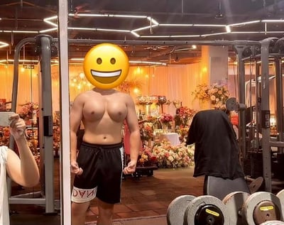 健身房场景中，一名赤裸上身的肌肉男正在自拍，其面部被黄色笑脸表情包覆盖（可能为保护隐私或增加趣味性）。画面左侧可见持手机自拍的手臂，背景包含杠铃架、哑铃等健身器材，同时意外出现大量鲜花装饰，形成硬汉健身场景与柔美花卉的强烈视觉反差。灯光明亮柔和，整体氛围诙谐又略带违和感。