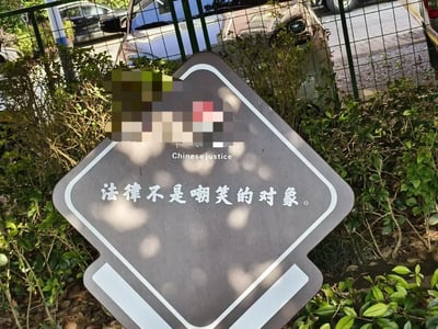 图片展示了一块灰色的警示牌，上面用白色字体写着“法律不是嘲笑的对象。”，下方还有英文翻译“Chinese justice”。牌子顶部有一块区域被马赛克处理。警示牌周围有绿色植物环绕，背景中可以看到铁丝网围栏和停放的汽车，整体场景像是在某个停车场或园区的户外区域。