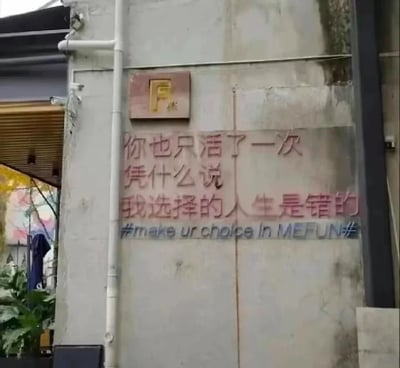 这是一张原始拍摄的静态标语图，画面主体是一面带有污渍和岁月痕迹的水泥墙面，墙面上印着醒目的红色中文标语：“你也只活了一次 凭什么说 我选择的人生是错的”，标语下方配有蓝色的英文话题标签“#make ur choice in MEFUN#”，墙面上方还有一个棕色底、黄色“F”字样的方形标识。画面左侧可见白色垂直排水管，排水管旁有绿色植物、收起的户外遮阳伞，以及部分带有彩色涂鸦的建筑结构，整体场景偏向街头文艺风格，传递出拒绝他人评判、坚持自我人生选择的鲜明态度。