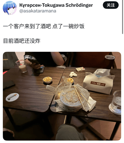 社交媒体用户发布的图文内容，文字部分描述“一个客户来到了酒吧 点了一碗炒饭 目前酒吧还没炸”，配图为酒吧内的餐桌实景。桌上有外卖炒饭（带筷子）、标有“chōu 抽我 wǒ”的纸巾盒、玻璃杯饮品、小碟坚果及正在使用手机的人，整体营造出一种日常中略带幽默的场景，调侃在酒吧点炒饭这一不太常见的行为。