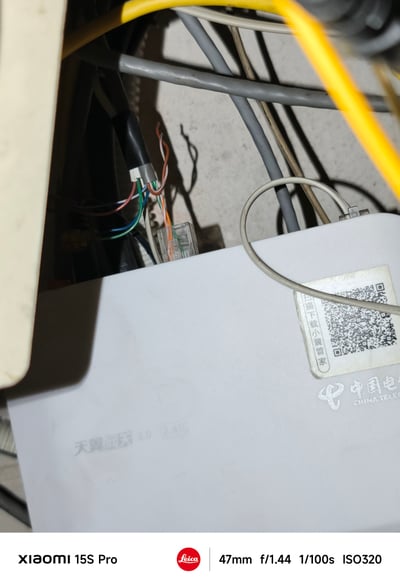 一张近距离拍摄的家庭网络设备特写照片，画面中心是中国电信的白色光猫设备，表面印有「天翼宽带3.0」字样及中国电信LOGO。设备顶部贴有带二维码的标签，标签上有「扫描下载小翼管家」字样。光猫周围缠绕着多根杂乱的线缆，包括黄色、灰色、黑色等不同颜色的网线和电源线，背景为水泥墙面，整体呈现出典型家庭网络布线的凌乱状态。照片底部显示拍摄设备为小米15S Pro手机，带有Leica标识及相机参数。