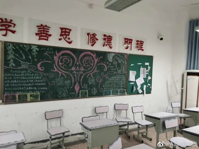 这是一张中学教室的实拍照片，教室墙面上方张贴着红色的“学 善思 修德 明理”德育标语；下方的大黑板布置了新学期主题黑板报，黑板报以粉色华丽对称花纹为中心装饰，左右两侧配有白色粉笔书写的文字内容与手绘图案，左侧是树木粉笔画，右侧是神情忧郁的动漫风格人物手绘，黑板上醒目写着“新学期 新气象”的字样，底部还有几个小的粉笔绘制的装饰框；右侧的小绿板上贴着一些不规则的白色纸片；教室下方排列着多套浅灰色的课桌椅，墙面有轻微的使用痕迹，整体呈现出典型的校园新学期的氛围，充满学生们对新学期的期待与布置教室的用心。