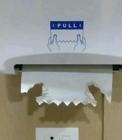 这是一张趣味恶搞的实拍图：公共抽纸机上方印有英文标识“PULL”和两只拉纸的简笔画手，下方的纸巾被撕出类似“嘴部咬住双手”的形状，和上方的简笔画形成视觉错位效果，仿佛是纸巾“咬住”了拉纸的手，制造出反差感满满的搞笑趣味，是利用日常物品制造的创意互动内容。