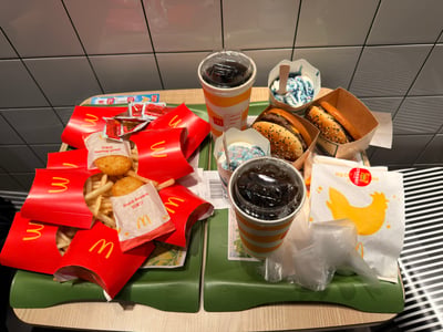 这张图拍摄于麦当劳门店内，浅木色桌面上摆放着两个绿色的麦当劳专用托盘：左侧托盘堆叠了多份红色包装的麦当劳薯条，还有两袋独立包装的脆薯饼，旁边放有数包番茄酱；右侧托盘有两杯带盖的加冰可乐、两个带芝麻面包的汉堡、两份表面带有蓝白色酱料的麦旋风冰淇淋，还有一份印有黄色小鸡图案的鸡肉类产品包装，背景是麦当劳门店常见的浅灰色方格瓷砖墙面。发帖人配文“薯条only”和实际满桌包含薯条、汉堡、饮品、甜品、小食的丰富餐品形成强烈反差，颇具调侃趣味。