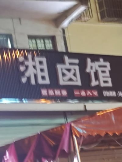 这是一张实地拍摄的街边湘卤馆店铺招牌，招牌采用黑底白字的设计，醒目的“湘卤馆”三个大字十分显眼，下方配有红色小字标语“百味鲜香 一卤入心”，右侧还有部分被截断的联系电话。招牌背景是居民楼建筑，下方可见红色的遮阳篷，整体呈现出普通街边餐饮小店的日常场景，没有双关、谐音梗等特殊内容。