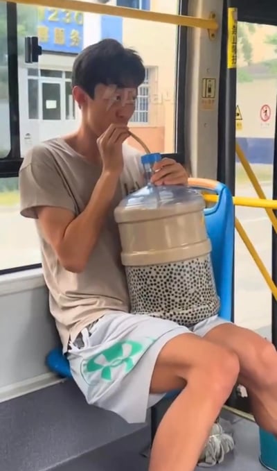 在公交车内的场景中，一名穿着浅灰色T恤的男生坐在座位上，通过视觉错位将大桶装饮用水放在腿与座椅之间，手持吸管做出喝水的动作，营造出抱着超大水桶喝水的搞笑假象，画面充满反差感和趣味性，让人看了忍俊不禁。