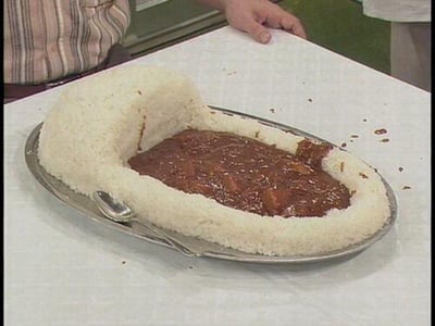 这张图片展示了一份设计十分新奇的料理：在金属餐盘上，白米饭被塑造成了餐盘的形状，围出一个凹槽，凹槽中盛放着深棕色的浓稠咖喱，旁边还摆放着一把金属勺子。背景中能看到两个人的部分躯体，暗示这是在一个节目场景中。这份料理打破了常规咖喱饭的呈现方式，用米饭模拟餐盘容器，创意十足，充满趣味性。