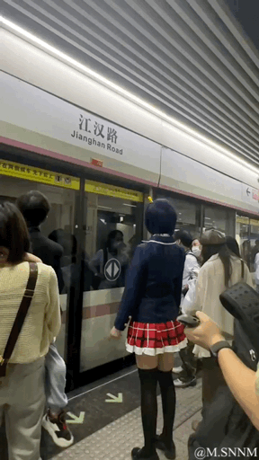 这是一段在武汉地铁江汉路站拍摄的动态画面，画面中一位身着蓝白制服上衣、红格短裙搭配黑色长筒袜的coser站在站台等候列车，身旁是众多普通候车乘客。地铁车门打开后，乘客们有序乘车，日常的通勤场景因为coser的出现增添了一丝新奇感，整体氛围平和自然。