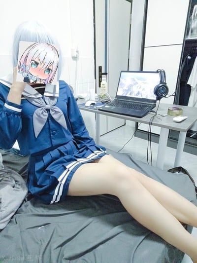 画面里是一位身着蓝灰配色JK制服的人，坐在铺有灰色床品的床上，手持一张带有害羞表情动漫少女的贴纸遮挡住脸部，贴纸配文“小哥一根不够！”。背景是一个摆放着笔记本电脑、头戴式耳机、鼠标的简易桌面，整体是日常居家的房间环境，风格轻松俏皮。
贴纸文字是带有玩笑性质的网络风格文案，无特殊双关或谐音梗。