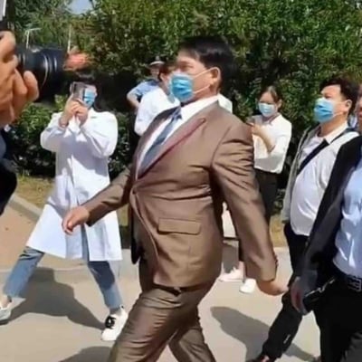 这是一张户外拍摄的静态纪实照片，画面中的所有人员都佩戴着蓝色口罩。中心位置是一位身着棕色西装、系着领带的男士，他正迈步前行，处于人群的核心位置，姿态正式。周围环绕着多位随行人员，其中有人手持专业相机、有人用手机进行拍摄记录，还有身着白色工作服的人员，背景是茂密的绿色灌木丛，整体场景氛围正式，看起来像是一次公务视察或者重要人物到访的活动现场，随行人员正在对该场景进行记录。