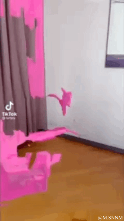 这是一段利用视觉错位效果制作的动态GIF。画面展示了一个室内场景，地板和墙面带有粉色色块，一把棕色椅子放置在地板的粉色区域上，同时空中有一个粉色的形状。从特定视角观察时，这些元素会组合成类似粉色动物（如马）的视觉假象，随着视角的轻微变化，这个假象会被打破，让人发现这只是场景布置形成的错觉，整体效果荒诞又充满趣味，给人新奇的视觉体验。