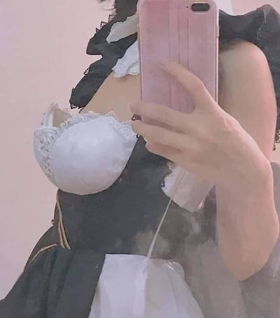 一位穿着女仆装的女性正在自拍，她手持粉色手机，手机挡住了脸部。上半身穿着白色蕾丝装饰的女仆装上衣，搭配黑色的女仆装元素，背景是纯色的浅粉色。