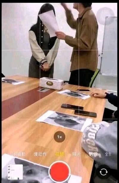这是一张竖屏手机格式的视频截图，画面呈现的是一个会议室场景：木质会议桌上摆放着多张印有同款怼脸表情包的纸张、几部智能手机以及一盒抽纸。画面中有三位真实人物，其中一位身着棕色上衣的人手持一张白纸，抬手做出要挥打向对面女生的动作；对面穿拼色棒球服外套的女生双手交握于身前，姿态显得拘谨。画面下方是手机相机的录制界面，红色录制按钮显示正处于视频拍摄状态，界面上有“视频”“慢动作”“照片”等拍摄模式选项。整个场景并非真实的指责或惩罚行为，而是职场同事之间的整活搞笑互动，是工作之余的趣味放松时刻。