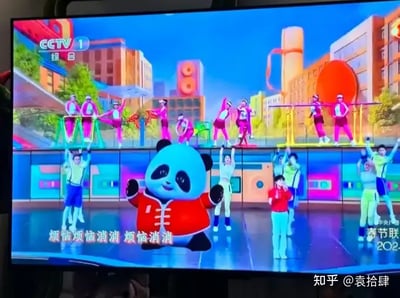 这是一张央视春晚晚会的电视截图，画面中央是一只穿着红色唐装的熊猫吉祥物，身边有众多跳舞的演员，整体色彩明亮活泼，背景采用卡通元素和现代建筑结合，氛围非常欢快。下方有中文歌词“烦恼烦恼消消消 烦恼消消”，表达了积极向上的节日主旨。画面没有明显双关、谐音梗或暗示内容，极具合家欢气质。