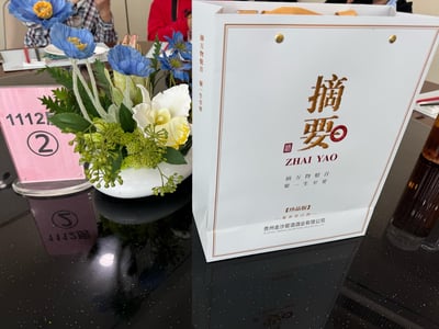 这是一张餐厅餐桌的实拍图，画面主体右侧是贵州金沙窖酒酒业出品的「摘要酒（珍品版）」白色手提礼品袋，袋身印有金色的「摘要」字样、「摘万物魁首 聚一生至要」的宣传语，明确标注为酱香型白酒；画面左侧是摆放着蓝色仿真花的桌面花艺装饰，旁边立着粉色的「1112房 ②」桌牌，背景中能看到餐厅餐具和正在用餐的人，整体呈现出商务聚餐的正式场景。