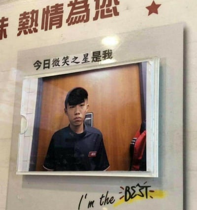 这是一张充满反差感的搞笑梗图：在一家标注着「熱情為您」的门店中，一个被选为「今日微笑之星」的员工，却摆出了一张严肃、满脸不情愿的臭脸，和上方的「今日微笑之星是我」以及下方的「I'm the BEST」形成强烈的视觉和语义反差，精准戳中打工人被迫营业、身不由己的情绪，引发共鸣。
没有双关、谐音梗，核心笑点来自标语与实际表情的巨大反差。