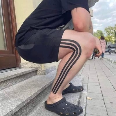 这是一张真人实拍的静态照片，画面主体是一名蹲在建筑户外石阶上的男子，他身着黑色短袖上衣、黑色运动短裤，脚穿黑色洞洞鞋。其右大腿到小腿的位置纹有三条平行的黑色长条纹，条纹顺着腿部的曲线自然延伸，在他蹲下的姿态下，纹身的上端刚好和短裤的裤边衔接，视觉上仿佛他穿着侧边带有经典三道杠设计的阿迪达斯运动长裤，形成了有趣的视觉错觉。男子右手握着一个金属喷头类的物品，背景可见城市人行道、远处的行人和树木，整体是日常的户外街景。