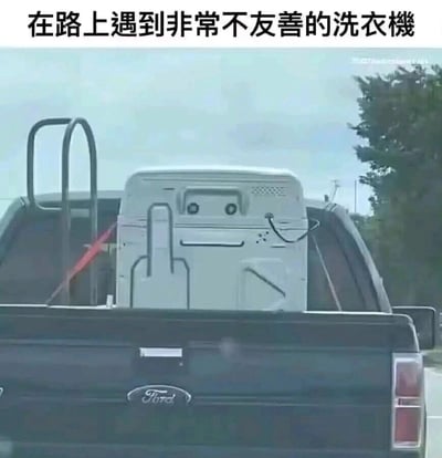 这是一张在道路上拍摄的福特皮卡尾部的照片，皮卡货厢内固定着一台洗衣机，洗衣机背部的结构、电线以及固定孔洞恰好组合成了类似生气皱眉的人脸图案，形成了有趣的视觉错觉。图片上方配有文字“在路上遇到非常不友善的洗衣机”，用拟人化的方式调侃这个巧合，让原本普通的搬运场景变得充满趣味性，属于生活中偶然捕捉到的趣味瞬间。
