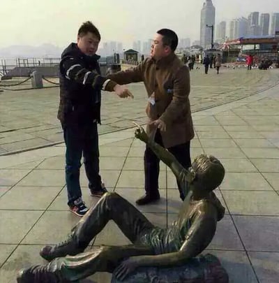 图片展现了两个路人在雕塑旁互动的有趣场景。一个路人和同伴一起指着地上的雕像，雕像本身也抬手指着路人，三者产生了一种互相推诿或指认的幽默气氛。整个场景发生在海边广场，远处有高楼大厦，环境显然是一个城市滨海区域。本图常用于讽刺生活中互相推卸责任、“甩锅”或是路人冷漠围观但不施以援手的现象。

- 本图产生了明显的双关感；既可解读为互相指责，也有讽刺人们在公共场合遇到问题时的冷漠。