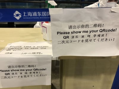 这是一张拍摄于上海浦东国际机场的现场实拍照片，展示了机场查验点位的工作场景。画面中的工作台上张贴了多张提示纸张，其中主要的提示内容以简体中文、英文、韩文、日文四种语言标注，要求旅客出示二维码；另有一张提示纸告知旅客需要截图保存二维码，同样包含英文和简体中文两种语言。画面右上角可见一位身着白色防护服、佩戴口罩的机场查验工作人员，背景处的蓝色标识牌上印有“上海浦东国际 SHANGHAI PUDONG INTERNATION”的字样，明确了拍摄地点。整个场景是机场查验流程的一部分，通过多语言提示满足不同国家和地区旅客的理解需求，引导旅客配合完成相关查验步骤。