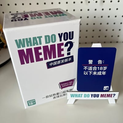 图片展示了What Do You Meme?桌游的中国首发版本。画面中央是一个白色游戏盒，上面印有彩色的"WHAT DO YOU MEME?"标题，下方有"中国首发版本"字样。盒子侧面标注了"亚马逊NO.1桌游 热卖全球70国"以及"AGES 18+"的年龄限制。盒子旁边立着一个蓝色的警告卡片，上面写着"警告：不适合18岁以下未成年"。整个产品放置在白色的展示台上，背景是带有圆孔的白色展板。
