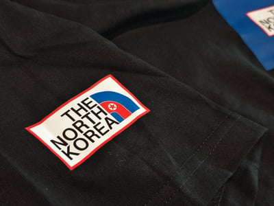 图片展示了一件黑色衣物的特写，上面有一个模仿The North Face品牌的恶搞logo补丁。补丁设计为红白蓝三色，中央文字为"THE NORTH KOREA"，并包含类似朝鲜国旗的元素。背景中还能看到另一个类似的补丁部分露出。这是对知名户外品牌logo的幽默改编，将"Face"替换为"Korea"并融入朝鲜国旗元素。