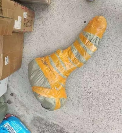 图片展示了一个放置在灰色水泥地面上的包裹场景。中央是一个被黄色胶带和灰色编织袋包裹的不规则形状物体，胶带呈横向缠绕，形成黄灰相间的条纹。左侧堆放着几个棕色纸箱，部分纸箱上有模糊的标签贴纸，右下角可见一个蓝色塑料袋的边缘。整体场景像是物流仓库、搬家现场或临时储物区域，物体可能是大型物品的包装状态。