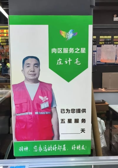 这是一张在超市内拍摄的「肉区服务之星」立牌照片，立牌上展示了名为庄计毛的工作人员的微笑照片，上方印有品牌标识「羽神」，标注了「肉区服务之星 庄计毛」的字样，下方写着「已为您提供五星服务____天」，但天数的填写位置是空白的，底部还有品牌标语「羽神，您永远的好邻居，好朋友！」。空白的天数栏形成了趣味点，仿佛还未开始统计服务天数或是遗漏填写，显得十分滑稽。